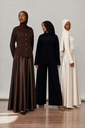 Three modesl wearing hijabs and long dresses in brown, black, and white against a plain background.